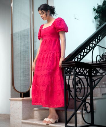 Model wearing a raspberry red eyelet cotton maxi dress with puff sleeves, square neckline, and tiered skirt, styled in a minimal indoor setting.
