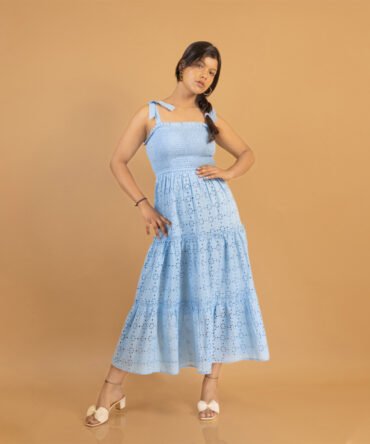 A woman wearing a blue cotton eyelet tiered maxi dress with broad bow tie straps, smocked bodice, and flowy skirt, posing stylishly against a tan background.