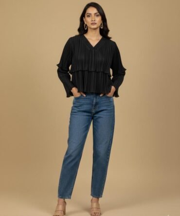 Woman wearing a black pleated ruffle top with flared sleeves and blue jeans, standing against a neutral beige background, minimal chic fashion look.
